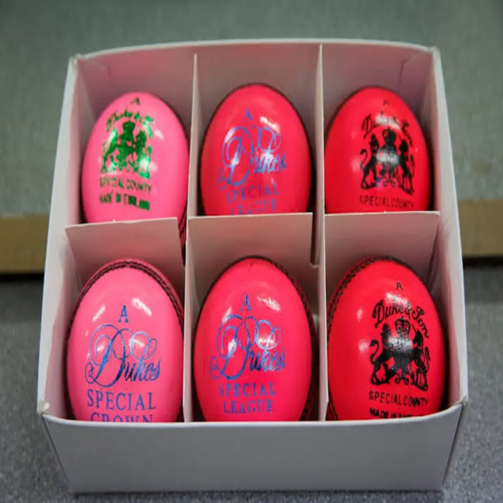 Six 11Sports high-visibility pink cricket balls with blue foil crests neatly boxed in cardboard dividers.