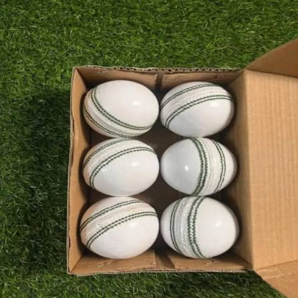 Cardboard box containing six 11Sports white leather cricket balls with green seam stitching on turf.