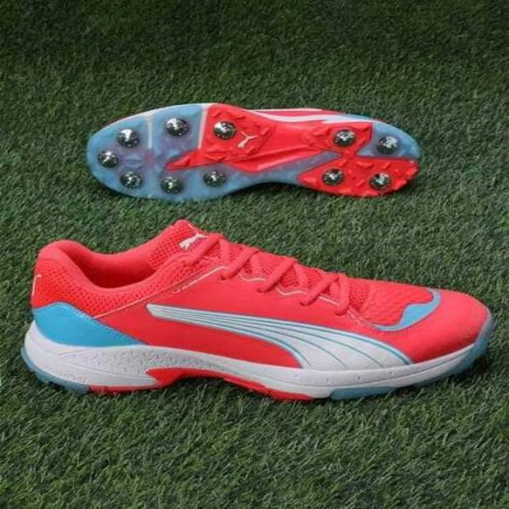 11Sports bright coral red cricket spikes with cyan blue accents resting on green turf beside its metal spike sole.